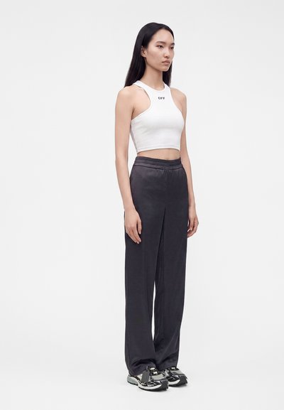 Femme aux longs cheveux noirs portant un débardeur court blanc, un pantalon noir ample et des baskets noir et blanc, debout devant un fond uni.
