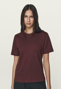 Bordowa koszulka z krótkim rękawem z miękkiej, gładkiej bawełny z klasycznym okrągłym dekoltem i luźnym fasonem, w połączeniu z czarnymi spodniami.