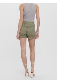 Pantaloni corti all'uncinetto verde oliva con orlo a scallop, caratterizzati da motivi intricati. Abbinati a una canotta grigia e sandali con tacco di colore chiaro.