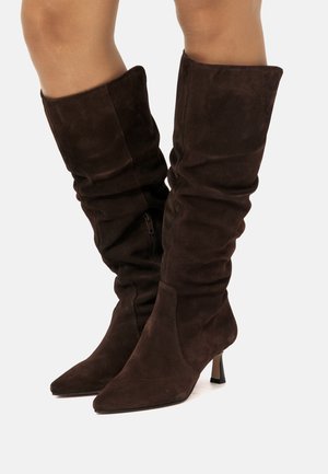 Botas altas de ante marrón con un diseño arrugado, puntera afilada y tacón cónico. Presentan un cierre lateral y una textura suave.