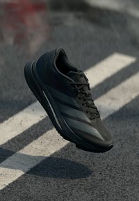 Chaussure de sport noire avec une tige en mesh texturé, des accents synthétiques lisses et une semelle en caoutchouc. Présente des bandes contrastantes gris foncé.