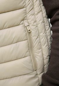 Beige Daunenweste mit gesteppten Abschnitten und einer seitlichen Reißverschlusstasche. Weiche, glatte Textur mit minimalen Details. Brauner Strukturstoff in der Nähe sichtbar.
