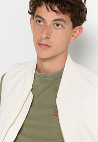 Lys beige bomberjakke med lynlås og ribbet krave, båret over en grøn crewneck trøje med broderet logo og små detaljer.