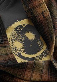 Pruunide ja kuldsete toonidega ruuduline välimine kiht, mis paljastab tumeda särgi, millel on kuldne naeratava näo graafika. Tekstuuriga kangas, millel on näha nööpide detail.