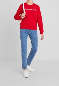 Sudadera roja con el logo "CALVIN KLEIN JEANS", combinada con jeans de mezclilla azul claro y zapatillas blancas. Estilo y diseño sencillos y casuales.