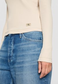 Beige Langarmoberteil mit CK-Logo-Tag, kombiniert mit blauen Jeans, zeigt einen Teil des Oberkörpers und die rechte Hand entspannt an der Seite.