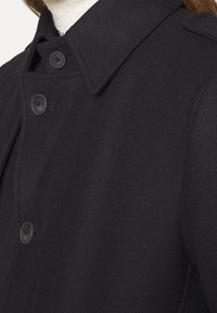 Cappotto in misto lana nero con chiusura a bottoni, colletto appuntito e tessuto testurizzato con un sottile motivo diagonale.