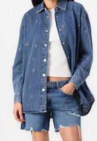 Džínová košile na knoflíky se stříbrnými švy, dlouhými rukávy a volným střihem, párovaná s opotřebovanými džínovými šortkami.