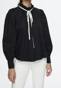 Zwarte blouse met pofmouwen, gerimpelde kraag en een contrasterende witte strik bij de halslijn. Gladdige stof, losse pasvorm, met een geplooid detail.