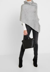 Femme portant un poncho en maille gris, un jean skinny noir, des bottes à talons aiguilles noires, et tenant un sac à main en cuir noir.