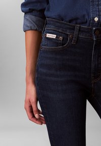 Blugi din denim albastru închis, cu talie înaltă, cusături cu fir cupru, buzunar frontal cu etichetă și închidere cu nasturi.