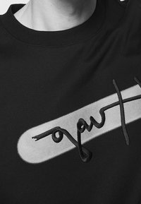 Camiseta negra de algodón con cuello redondo y un logo central en negrita sobre un parche gris curvado. Textura suave.