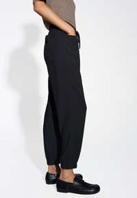 Pantaloni neri larghi con polsini elastici, tasche laterali e vita con cordino. Indossati con scarpe nere, il tessuto sembra liscio.