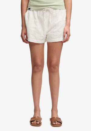 Kvinde iført hvide broderede shorts med snøre og lommer, lysegrøn top og brune slid-sandaler, stående mod ensfarvet baggrund.