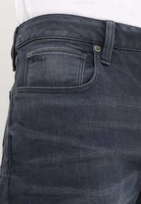 Jeans i mörkblå denim med en något blekt textur. Har en framficka och metallnitar. Syrader är synliga längs midjelen.