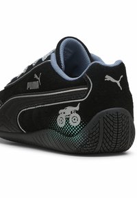 Črne semiš superge s srebrnim logotipom Puma, subtilna grafika monstrous kamiona in teksturiran črn gumijasti podplat z zelenim gradientnim detajlom.