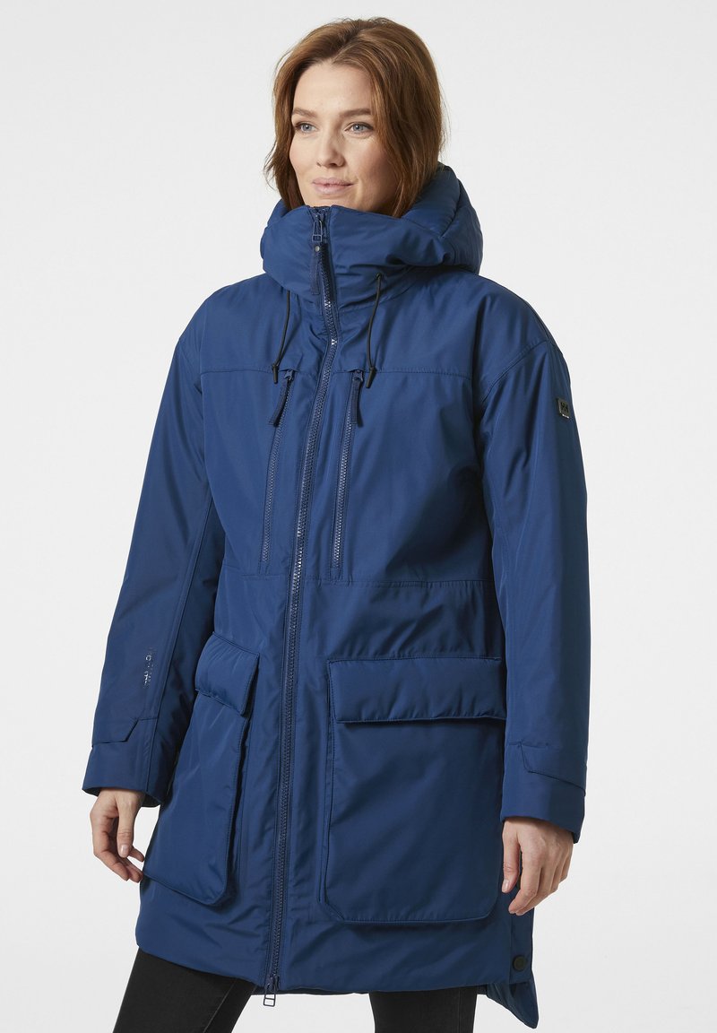 Un largo abrigo parka azul marino con un cuello alto, cierre de cremallera, capucha ajustable y grandes bolsillos frontales. Fabricado con un material suave y resistente a las inclemencias del tiempo.