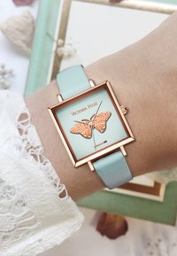 Montre carrée arborant un cadran bleu avec un motif de papillon, des détails en or rose et un bracelet en cuir bleu clair. Convient pour une tenue décontractée.