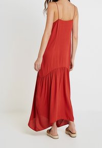 Maxi sukienka w kolorze czerwonym z regulowanymi ramiączkami, zwiewnym materiałem i ściągnięciem w talii. Dół jest dłuższy z tyłu o lekkim kształcie litery A.