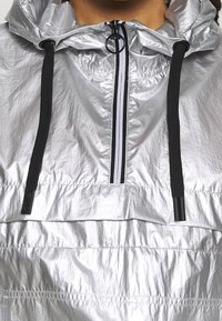 Silverfärgad huva vindjacka i glänsande material, med halvdragkedja fram, svarta dragskon och en texturerad yta.