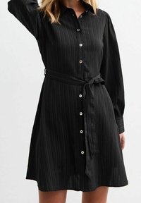 Abito camicia nero a maniche lunghe con bottoni frontali, vita con fiocco e texture a righe verticali. Presenta dettagli in bottoni argentati.