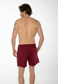 Bordeaux-värgade badshorts med elastisk midjeband, bakficka och slät textur, bärs av en person med tatueringar, sedd bakifrån.