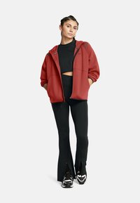 Felpa rossa con zip, dal taglio rilassato, abbinata a pantaloni neri a vita alta con cerniere sui lati e sneakers bianche e nere.