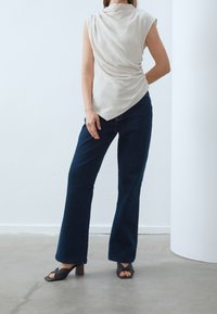 Haut beige sans manches avec dos drapé, associé à un jean large bleu foncé et des sandales à talons noires, sur fond neutre.