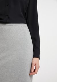 Harmaa puuvillainen kynähame, jossa on ribbipitsivartalo, yhdistetty mustan pitkähihaisen paidan kanssa. Käsi lepää hameen päällä, osoittaen kankaan rakennetta.