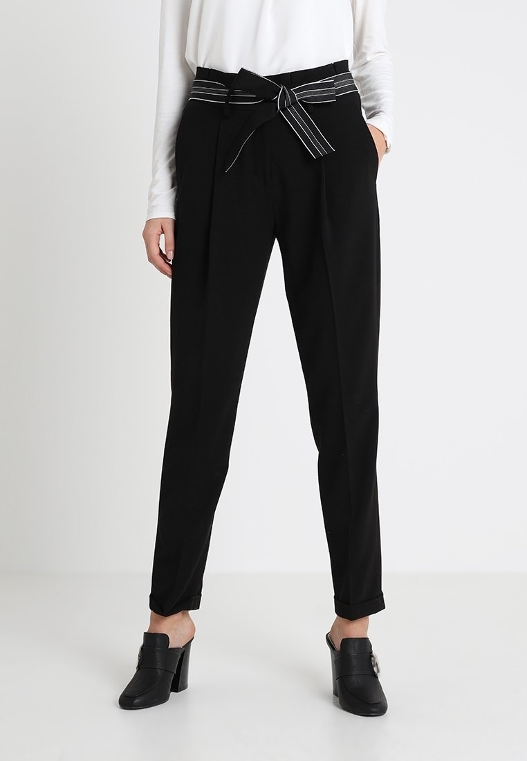 Femme portant un pantalon noir taille haute avec une ceinture en tissu rayée, un haut blanc à manches longues et des mules noires à talons, debout les mains dans les poches.