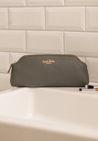 Grijze, gestructureerde leren make-uppouch met een gouden rits en branding op de rand van een witte wastafel, vergezeld van twee kleine make-upkwasten.