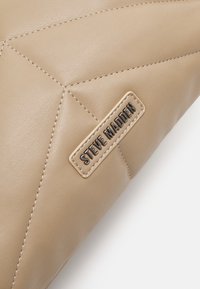 Róg beżowej pikowanej skórzanej torby z widocznym przeszyciem i naszytą metalową prostokątną etykietą z napisem "STEVE MADDEN."