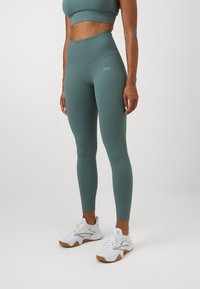 Colanți din material elastic, de culoare teal, cu talie înaltă și textură netedă, având un branding subtil, asortați cu pantofi sport albi și un top scurt în aceeași nuanță.