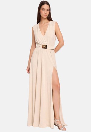 Donna che indossa un vestito maxi beige senza maniche con scollatura profonda a V, vita cintata, spacco sopra la coscia e tacchi argentati con cinturini, in piedi davanti a uno sfondo semplice.