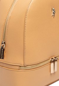 Sac à dos en cuir beige texturé avec des fermetures éclair et des accessoires dorés. Il présente une forme arrondie et un détail de logo sur le côté.
