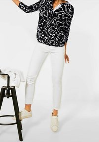 Kvinna i en blus med svart-vitt mönster, vita slim fit-byxor och vita sneakers, stående bredvid en svart pall med en vit skjorta.