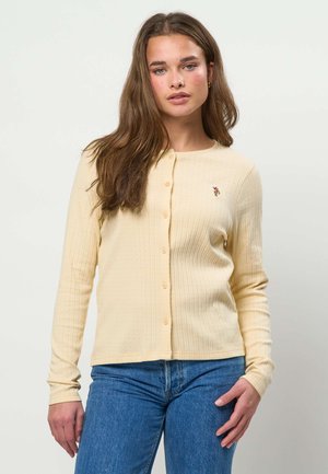 Noor naine pikkade pruunide juustega kannab helekollast nööbitavat kardigani ja siniseid teksaseid, ta seisab ühetaustalise valge tausta ees.