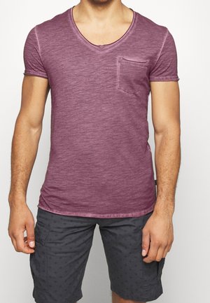 Homme portant un t-shirt ajusté à col en V bordeaux avec une poche poitrine et un short gris foncé à motifs aux genoux.