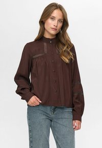 Vrouw met lang golvend haar, gekleed in een donkerbruine blouse met knopen en kanten inzetstukken, en een blauwe spijkerbroek, staande voor een effen achtergrond.
