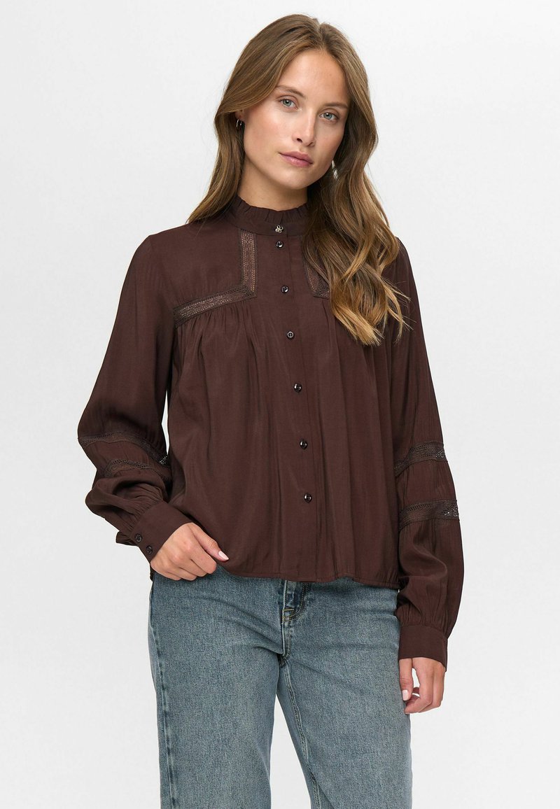 Vrouw met lang golvend haar, gekleed in een donkerbruine blouse met knopen en kanten inzetstukken, en een blauwe spijkerbroek, staande voor een effen achtergrond.
