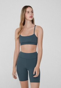 Sujeción deportiva en color verde azulado oscuro con tirantes finos, combinada con pantalones cortos de cintura alta a juego. Ambos están hechos de una tela suave y elástica.