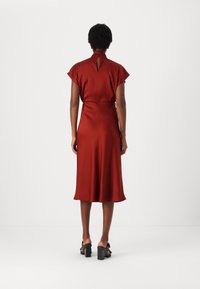 Abito in satin rosso con colletto alto, maniche corte, dettaglio a chiave sulla schiena e orlo svasato, abbinato a sandali neri con tacco a blocco e cinturini.