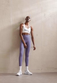 Balti Nike sportinė liemenėlė, levandų spalvos aukštai juosiai tamprės ir baltos kedai. Lygus audinys su subtiliais logotipo akcentais; minimalistiškas dizainas.