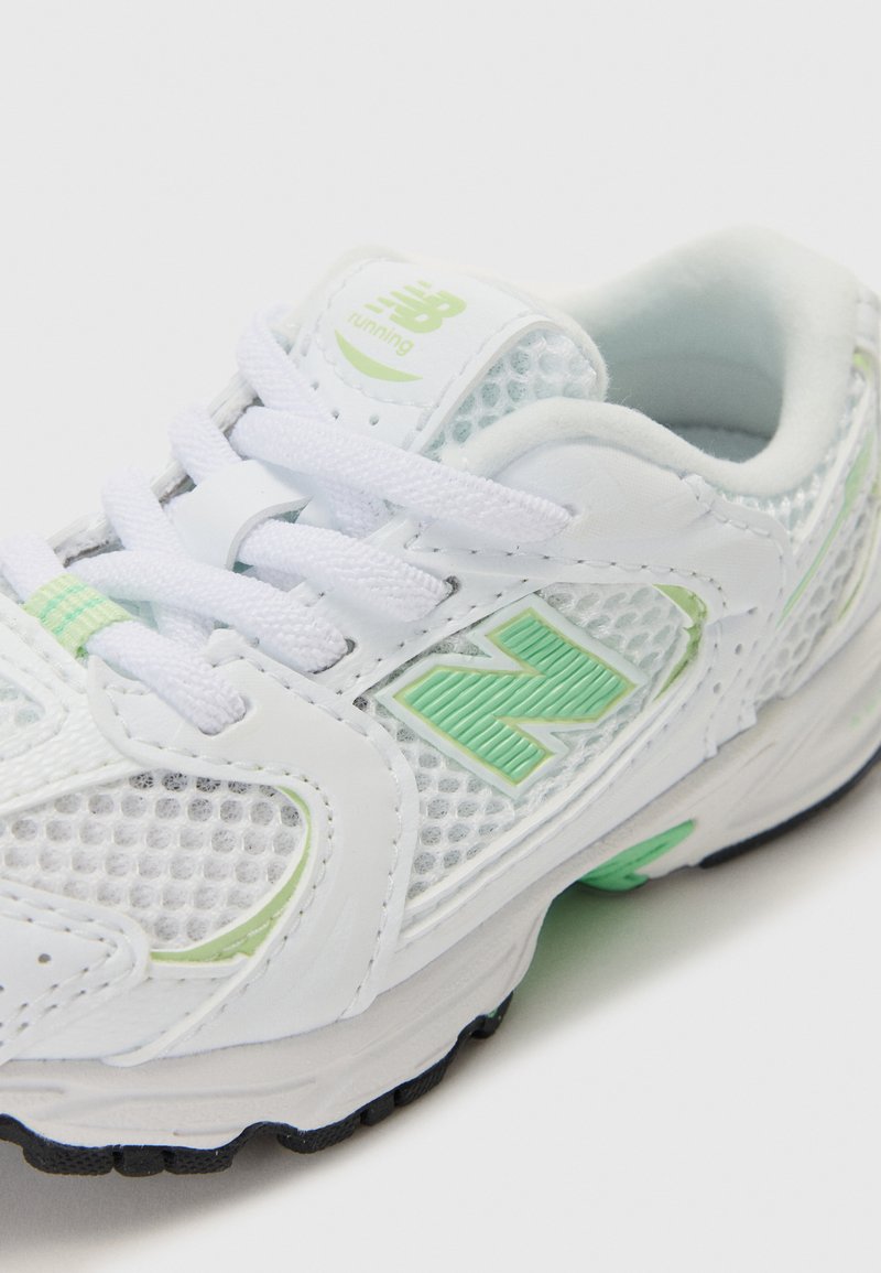 Bottine de sport blanche en matériau mesh, avec des accents verts et une semelle texturée. Comprend des lacets blancs et un logo sur la languette.