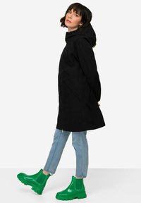 Imperméable noir avec capuche, doté de poches latérales et d'un accent de zip vert ; associé à des bottines vertes brillantes et à un jean bleu clair.