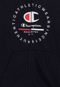 Felpa con zip color navy realizzata in tessuto morbido, con un logo circolare che presenta la scritta "Champion" in bianco e rosso, e tasche frontali.