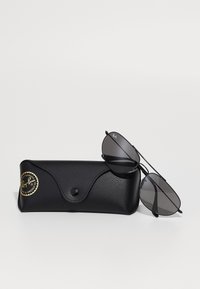 Occhiali da sole aviator Ray-Ban neri con lenti scure appoggiati su un astuccio in pelle nera con texture e un adesivo del logo dorato.