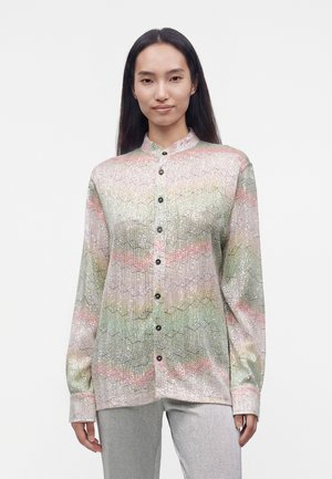 Kvinde med langt mørkt hår iført en pastel flerfarvet, mønstret skjorte med knapper og lysegrå bukser, stående mod en ensfarvet baggrund.