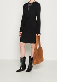 Abito lungo nero a maniche lunghe con cintura legata in vita, abbinato a stivali cowboy neri a punta e a una borsa a tracolla in suede marrone. Texture liscia in tutto l'abito.