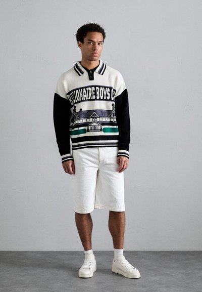 Pull en tricot avec des sections noires et crème, présentant un design graphique de maison et du texte. Associé à un short en denim blanc et à des sneakers blanches.